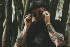 A fashionable man adjusting his sunglasses amidst a bamboo forest, showcasing tattoos and accessories.