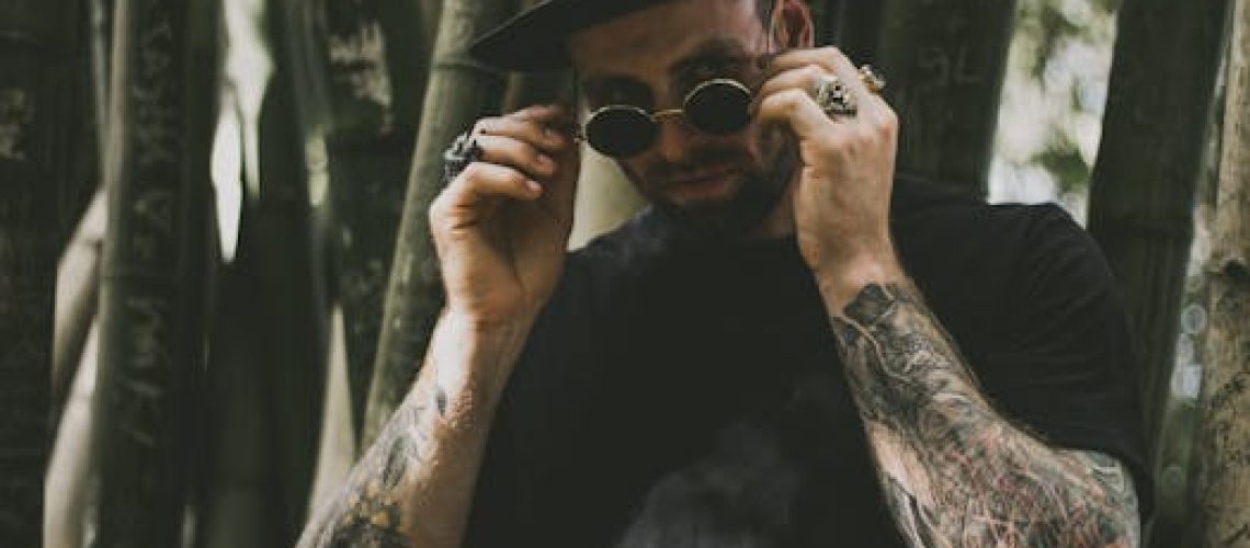 A fashionable man adjusting his sunglasses amidst a bamboo forest, showcasing tattoos and accessories.