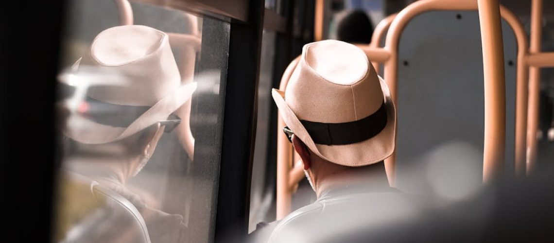 A man wearing a fedora hat sits inside a bus, reflecting on the window during a daytime commute.