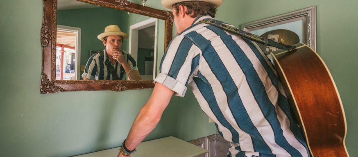 A musician in a fedora stands thoughtfully by a mirror with an acoustic guitar indoors.