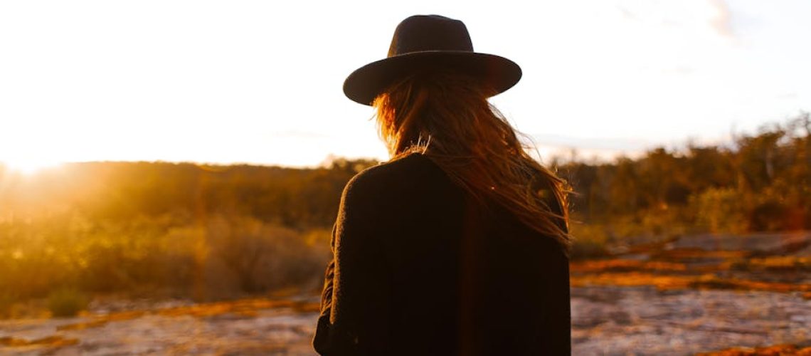 A woman in a fedora looks out over a sunlit countryside landscape during sunset.